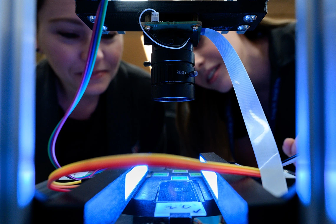 two people closely examining a piece of equipment under a camera setup