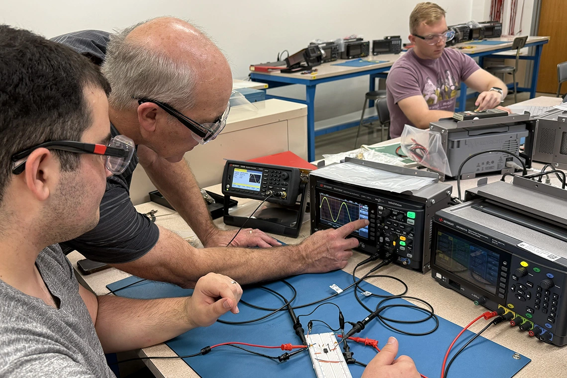 three people in electronics lab, two pointing to equipment
