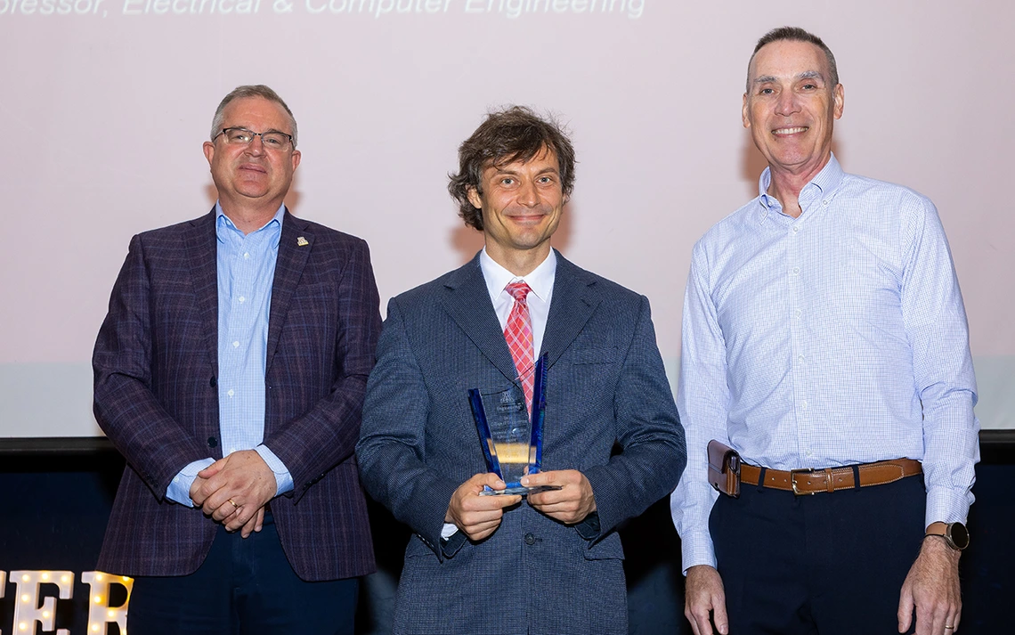 Three men pose for a photo. The man in the middle holds a glass award.