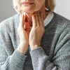 An older woman in a grey sweater touches her neck.