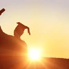 student in graduation regalia, holding up diploma, sunset in background
