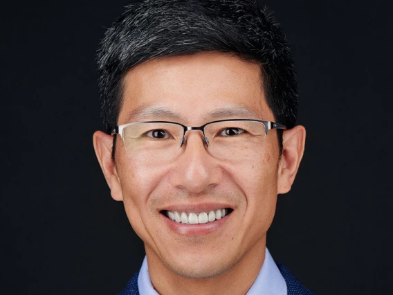 A man with glasses wears a blue suit and stands in front of a dark background.