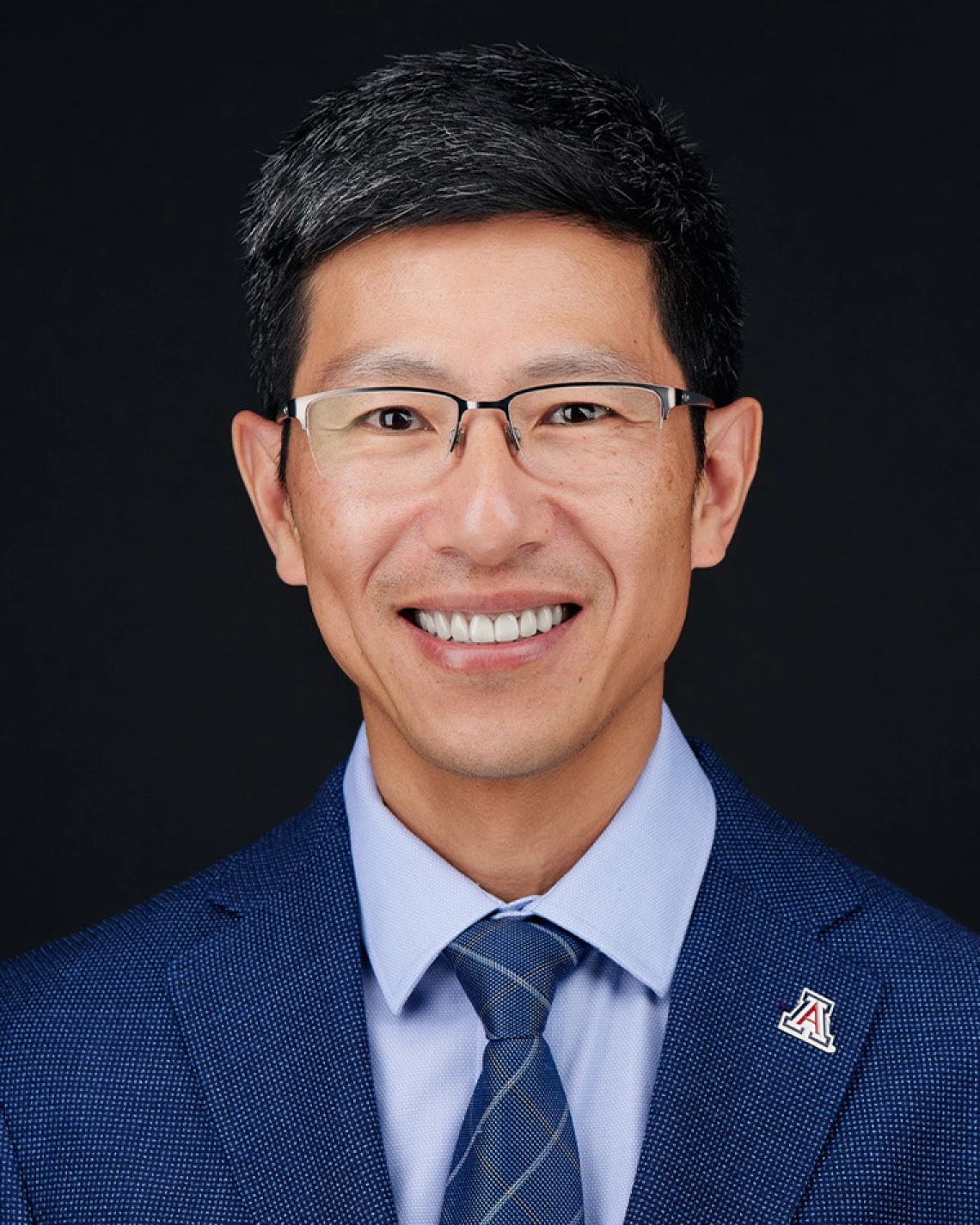 A man with glasses wears a blue suit and stands in front of a dark background.