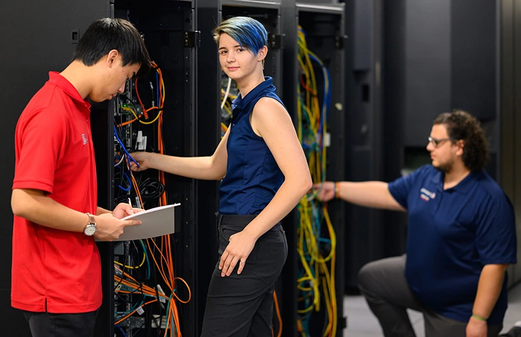 three people, two standing, one kneeling next to computer equipment