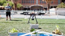 A drone floats over a green lawn. A man in the background controls the drone.