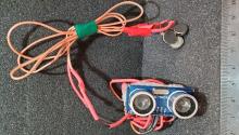 a boxy device a few inches wide with two round sensors. it looks similar to a pair of binoculars and a set of orange and red wires are attached.