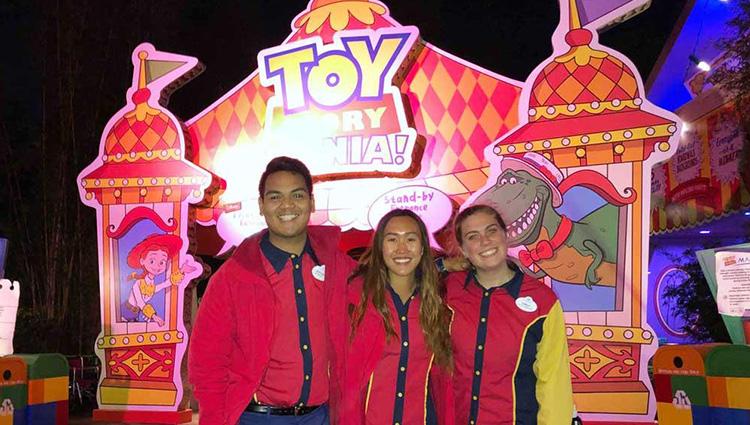Three smiling people in Disney uniforms stand in front of the Toy Story Mania! attraction.